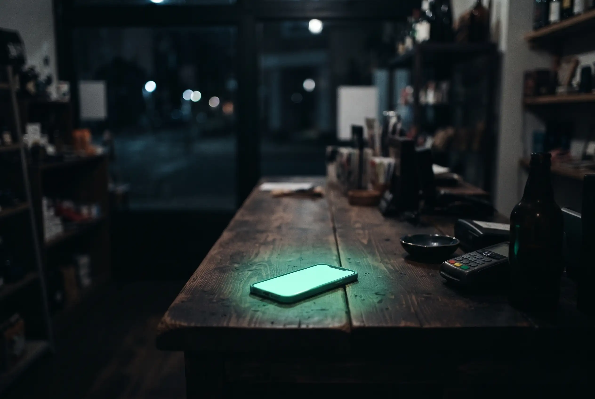 A shop counter after dark, a phone lying face up with its screen glowing, a card reader beside it.